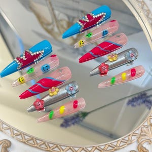 Op de afbeelding: Een set van tien kunstnagels met verschillende ontwerpen. De nagels zijn lang en puntig, met een verscheidenheid aan kleuren en patronen, waaronder roze, blauw, zilver en rood. Sommige nagels hebben smiley-gezichten, bloemen en glitter.