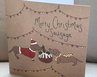 May include: A folded Christmas card with a brown kraft paper texture. The card features a hand-drawn illustration of two dachshunds, a Christmas pudding, and string lights. The text reads "Merry Christmas Sausage".