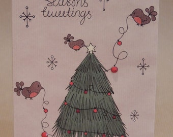 May include: A Christmas card with a hand-drawn illustration. The card features a green Christmas tree decorated with red ornaments, brown birds, and the words "seasons greetings". Presents are at the base of the tree, and snowflakes are scattered around.