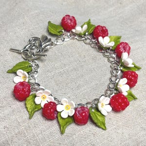 May include: A silver-toned chain bracelet featuring a series of red raspberry charms, white five-petalled flowers with yellow centres, and green leaves. The bracelet is secured with a silver-toned toggle clasp featuring a fleur-de-lis design.
