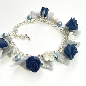 Peut inclure: Un bracelet à chaîne argentée orné de breloques en forme de roses bleu marine, d'accents de feuilles argentées et de petites perles bleues et blanches. Le bracelet a un fermoir mousqueton.