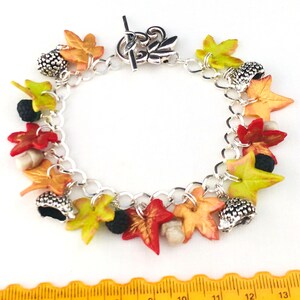 Puede incluir: Un brazalete de plata con dijes de tem&aacute;tica oto&ntilde;al, incluyendo hojas en tonos rojos, naranjas, amarillos y verdes, y dijes de bellota.