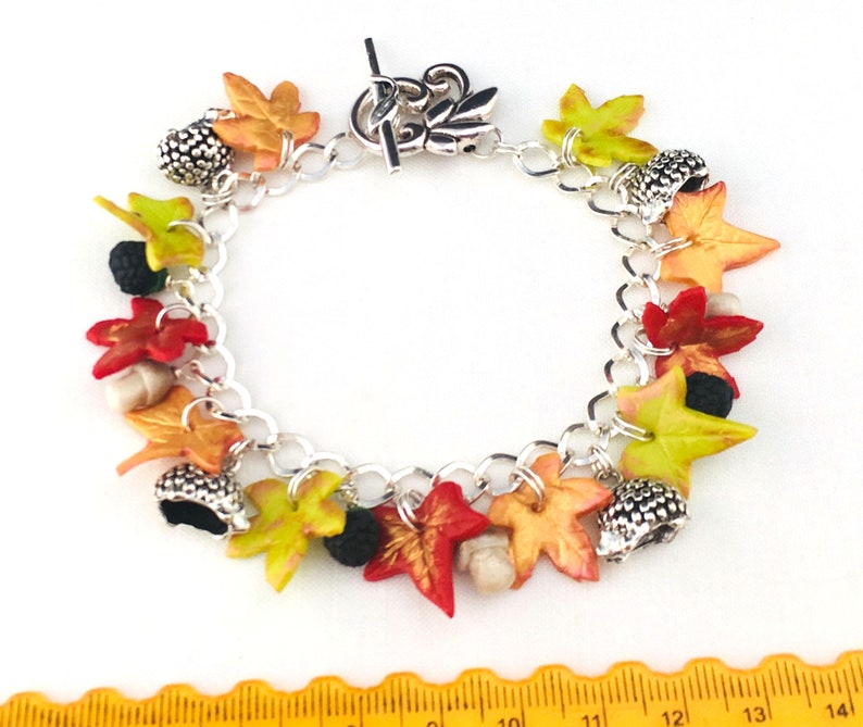 Puede incluir: Un brazalete de plata con dijes de tem&aacute;tica oto&ntilde;al, incluyendo hojas en tonos rojos, naranjas, amarillos y verdes, y dijes de bellota.