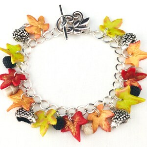 Puede incluir: Un brazalete de plata con dijes de tem&aacute;tica oto&ntilde;al, incluyendo hojas en tonos rojos, naranjas, amarillos y verdes, bellotas y moras.