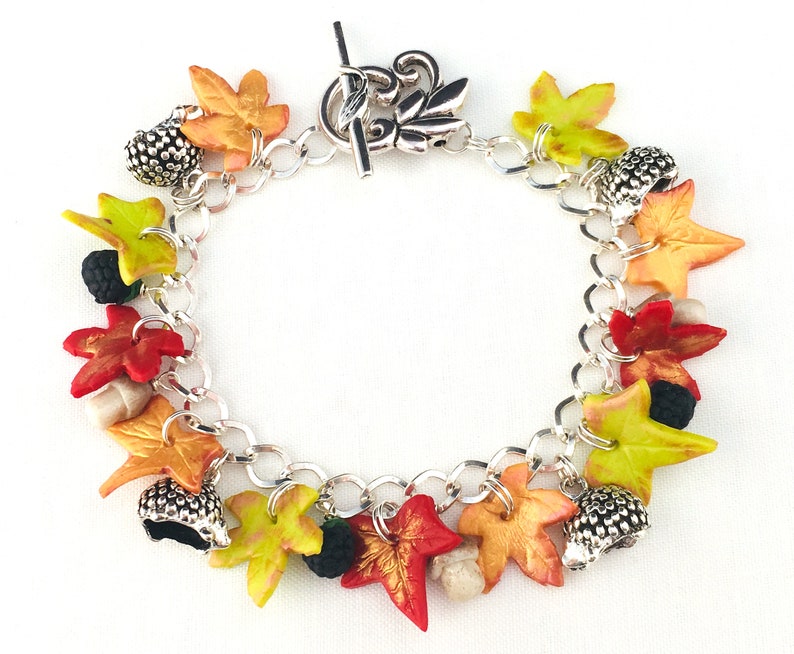 Puede incluir: Un brazalete de plata con dijes de tem&aacute;tica oto&ntilde;al, incluyendo hojas en tonos rojos, naranjas, amarillos y verdes, bellotas y moras.