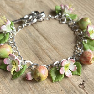 May include: A silver chain bracelet with delicate charms of pink and white flowers and green leaves. The charms are made of polymer clay and resemble miniature apples.