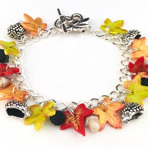 Puede incluir: Un brazalete de plata con dijes de tem&aacute;tica oto&ntilde;al, incluyendo hojas en tonos rojos, naranjas y amarillos, bellotas y moras.