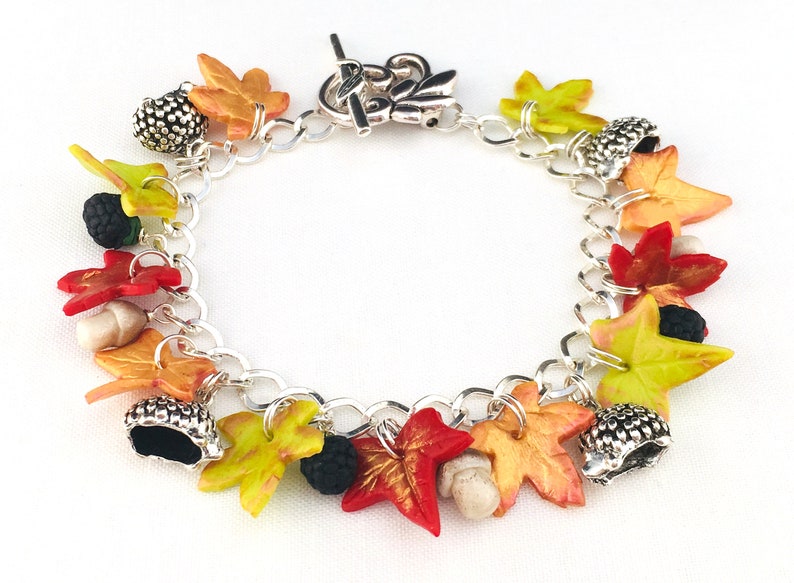 Puede incluir: Un brazalete de plata con dijes de tem&aacute;tica oto&ntilde;al, incluyendo hojas en tonos rojos, naranjas y amarillos, bellotas y moras.