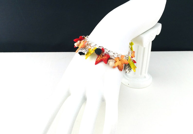 Puede incluir: Un brazalete de plata con dijes de tem&aacute;tica oto&ntilde;al. Los dijes est&aacute;n hechos de arcilla polim&eacute;rica y presentan hojas en tonos rojos, naranjas, amarillos y verdes. Tambi&eacute;n hay peque&ntilde;os dijes de bellota.