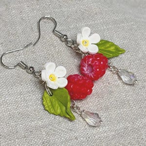 May include: A pair of dangle earrings featuring red raspberry charms, white five-petal flowers with yellow centers, green leaves, and clear faceted teardrop beads suspended from silver-toned metal hooks on a textured light gray fabric background.