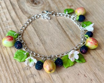 May include: A silver-colored chain bracelet adorned with miniature charms. The charms include green leaves, white flowers, and small fruits in shades of red, yellow, and black. The bracelet is set against a wooden background.