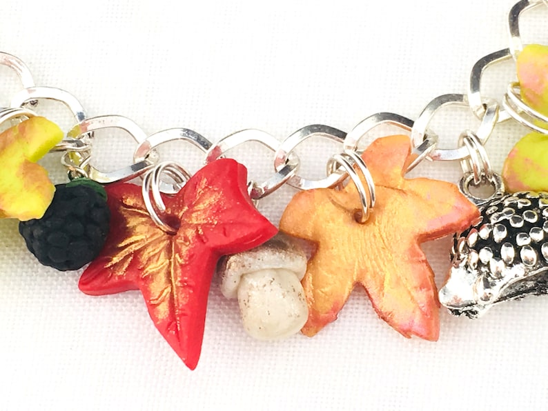 Puede incluir: Un brazalete de plata con dijes de tem&aacute;tica oto&ntilde;al, que incluyen una hoja roja, una hoja dorada, un hongo, una mora y un erizo.