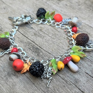 May include: A silver charm bracelet featuring a range of charms, including mushrooms, berries, leaves, and a fairy. The charms are crafted from polymer clay and painted in realistic colours.