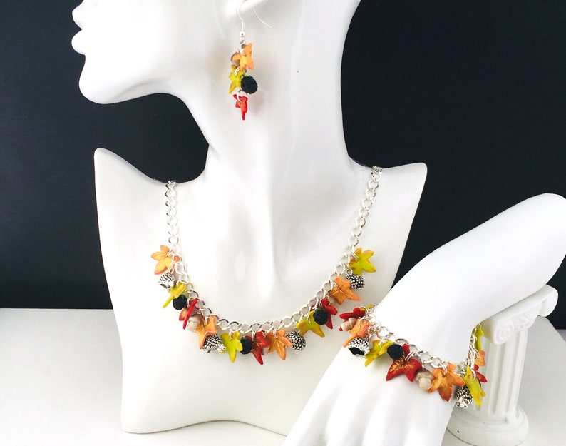 Puede incluir: Un collar y una pulsera de cadena plateada con dijes de tem&aacute;tica oto&ntilde;al. Los dijes est&aacute;n hechos de arcilla polim&eacute;rica y presentan hojas en tonos naranja, amarillo y verde, as&iacute; como peque&ntilde;as cuentas negras.