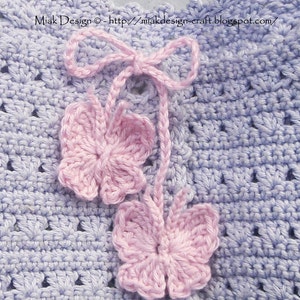 Peut inclure: Un tissu crocheté violet clair orné de deux papillons en fil rose suspendus à un nœud central. Le texte Miak Design - http://miakdesign-craft.blogspot.com/ est imprimé sur le haut du matériau texturé aux bords festonnés.