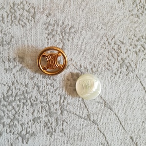 Op de afbeelding: Twee knopen met het Celine-logo. De ene knop is van goudkleurig metaal en de andere van wit plastic.