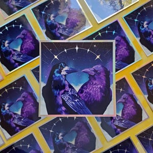 Puede incluir: Adhesivo cuadrado con acabado holográfico que representa dos cuervos morados frente a un cielo estrellado. El adhesivo está rodeado de otros adhesivos similares sobre una superficie amarilla.