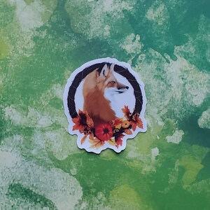 Puede incluir: Una pegatina con un zorro rojo con cara marrón y blanca, enmarcado en una corona oscura. La corona está adornada con hojas de otoño y una calabaza. La pegatina está sobre un fondo verde y blanco.