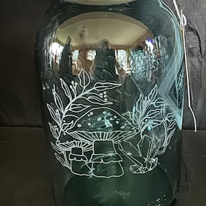 Könnte beinhalten: Ein grünes Glasgefäß mit einem silbernen Deckel und einem weißen, geätzten Design von Pilzen und Blättern. Das Gefäß ist mit "Made in USA" beschriftet.