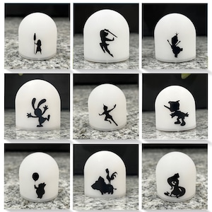 May include: Nine white dome-shaped night lights with black silhouettes of Disney characters. The characters include Peter Pan, Tinkerbell, a rabbit, a pig, a rooster, a bear, a mermaid, a princess, and a snowman.
