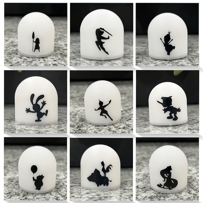 May include: Nine white dome-shaped night lights with black silhouettes of Disney characters. The characters include Peter Pan, Tinkerbell, a rabbit, a pig, a rooster, a bear, a mermaid, a princess, and a snowman.