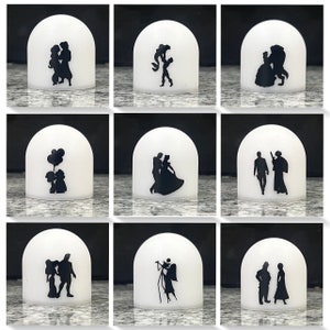 Könnte beinhalten: Neun weiße, kuppelförmige Kerzenhalter mit schwarzen Silhouetten von Disney-Paaren und -Charakteren. Die Silhouetten umfassen Belle und das Biest, Aschenputtel und den Prinzen, Arielle und Prinz Erik, Aladdin und Jasmin, Schneewittchen und den Prinzen, Rapunzel und Flynn Rider, Mickey Mouse und Minnie Mouse sowie Jack Skellington und Sally.