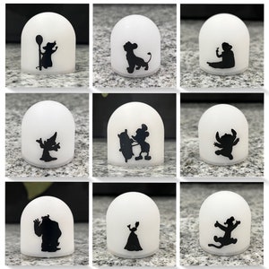 May include: Nine white dome-shaped night lights with black silhouette images of Disney characters. The characters include Mickey Mouse, Stitch, Simba, and more.