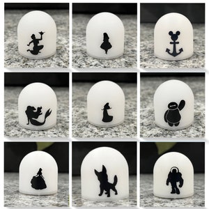May include: Nine white dome-shaped night lights with various black silhouette designs. The designs include Disney characters like Ariel, Cinderella, and Mickey Mouse, as well as characters from Pixar movies like Baymax and Dug.