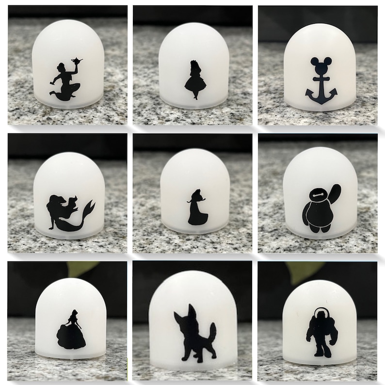May include: Nine white dome-shaped night lights with various black silhouette designs. The designs include Disney characters like Ariel, Cinderella, and Mickey Mouse, as well as characters from Pixar movies like Baymax and Dug.