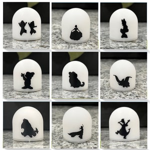 May include: Nine white night lights with different black silhouette designs. The designs include a princess, a dog, a dragon, a bear, a couple, a bird, a dinosaur, a character from the movie 'The Lion King', and a character from the movie 'Frozen'.