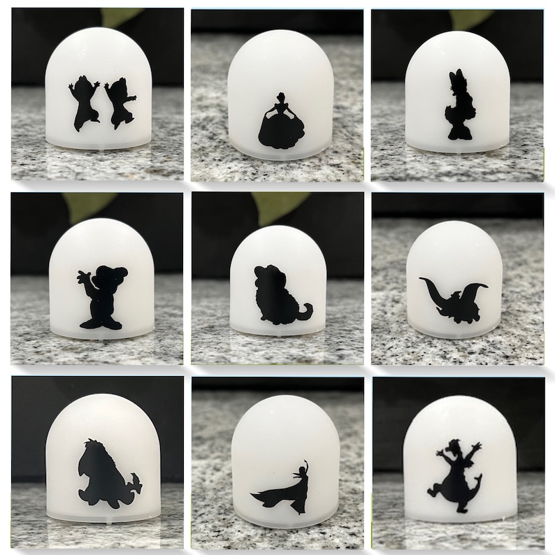 May include: Nine white night lights with different black silhouette designs. The designs include a princess, a dog, a dragon, a bear, a couple, a bird, a dinosaur, a character from the movie 'The Lion King', and a character from the movie 'Frozen'.