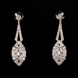 Peut inclure: Une paire de boucles d'oreilles pendantes de style Art Déco. Chaque boucle d'oreille présente un clou rond, un motif géométrique avec un diamant central et un pendentif orné pendant. Les boucles d'oreilles sont argentées et incrustées de nombreuses petites pierres scintillantes.