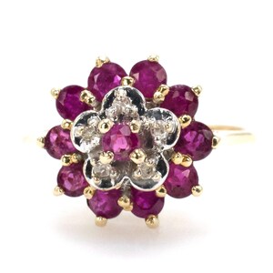 May include: A gold ring with a floral design. The ring features a central flower shape with small diamonds, encircled by a ring of deep red rubies. The ring's band is a thin, simple gold design. The gemstones are set in a gold setting.