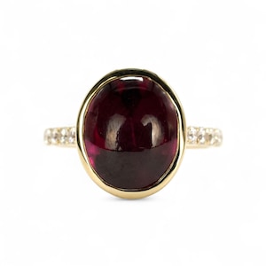 May include: A gold ring featuring a large, oval, deep red gemstone. The gemstone is set in a gold bezel. The ring band is adorned with small, clear gemstones. The ring is set against a white background.
