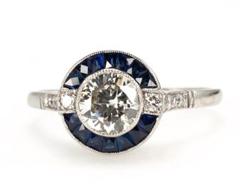 May include: A platinum ring featuring a large, round, brilliant-cut diamond center stone, surrounded by a halo of square-cut blue sapphires. The ring has additional small diamonds on the band, creating a vintage, art deco style.