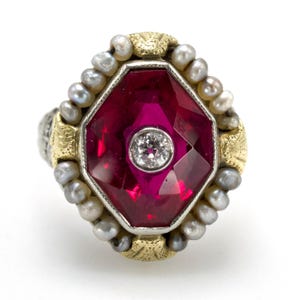 May include: A close-up of a ring featuring a large, faceted, ruby-red gemstone with a small diamond at its center. The ring is surrounded by small pearls and gold accents, set on a silver band. The ring has an antique or vintage style.