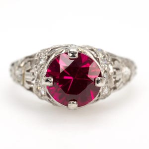 May include: A vintage-style ring with a large, round, deep red gemstone as the focal point. The setting is ornate, with intricate filigree and small accent stones. The ring is set against a white background.