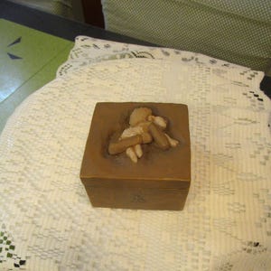 May include: A brown square box with a relief carving on the lid. The carving depicts a figure holding a smaller figure. The box is sitting on a white lace tablecloth.