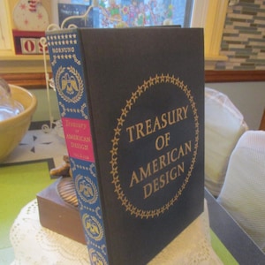 May include: A hardcover book titled "Treasury of American Design" with gold lettering on a dark blue cover. The spine is blue with gold accents and the title "Treasury of American Design" is printed on a red label. The book is displayed on a white lace doily.