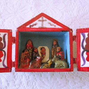 May include: A hand-painted, wooden nativity scene in a red box with opening doors. The box features a cross on top and decorative artwork on the doors. Inside, figures depict the traditional Christmas story.