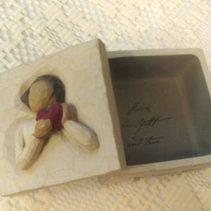 May include: A beige square box with a hinged lid, featuring a carved figure holding a heart. The interior of the box is dark, with the words "Love, beautiful, and true" inscribed. The box is open, revealing the inside.
