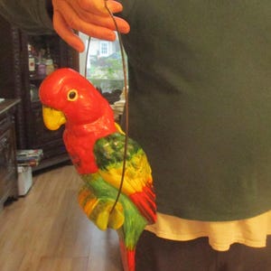 May include: A colorful ceramic parrot figurine with a red head, yellow beak, and green, yellow, and red body. The parrot is suspended by a brown string. The figurine is held by a person.