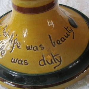 May include: A yellow ceramic base with a green and brown rim. The base has a handwritten inscription that reads "And that life was beauty was duty".