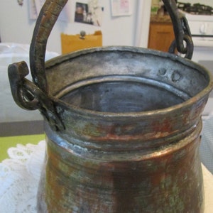 May include: An antique copper pot with a tarnished finish and a worn handle. The pot has a wide base and a rounded body.