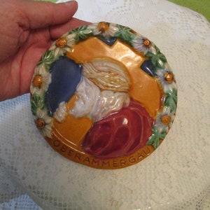 May include: A round ceramic plaque with a raised image of a person in a red robe. The plaque is decorated with a wreath of white flowers and green leaves. The words "Oberammergau" are written in brown lettering around the edge of the plaque.