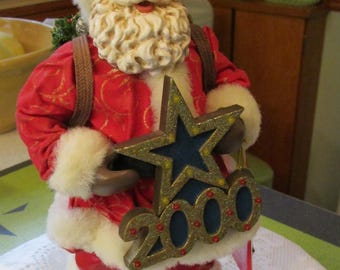 May include: A Santa Claus figurine wearing a red coat with gold swirl patterns and white faux fur trim. The figure has a white beard, a hat with a holly sprig, and black boots. It holds a gold star with the blue text 2000 and red accents.