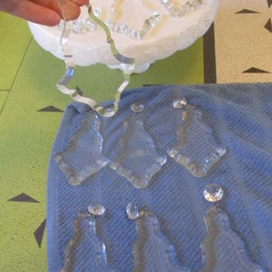 May include: Clear glass chandelier crystals with a faceted design and a small crystal at the top. One crystal is held by a hand, while others rest on a blue towel. A white, decorative object is in the background.