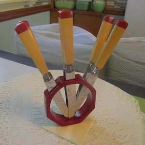 May include: A set of four steak knives with yellow handles and red tips, held upright in a red octagonal plastic stand. The knives feature metallic blades and are arranged in a circular pattern within the stand, which rests on a square base.