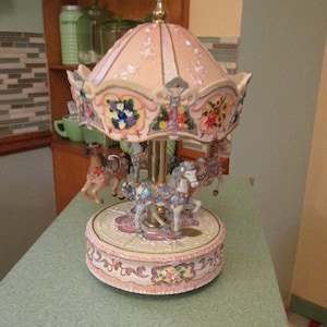 May include: A decorative carousel music box featuring a pale pink canopy with floral accents and gold trim. The base is circular with ornate scrollwork and floral details. Several miniature horses with intricate saddles and harnesses are suspended from the central gold-toned pole, rotating around the center.