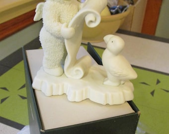 May include: A white porcelain figurine of a child in a hooded snowsuit with small wings holding a scroll, standing next to a small bird on a snowy base. The figurine is placed on a dark green box labeled Winter Tales of the Snowbabies.